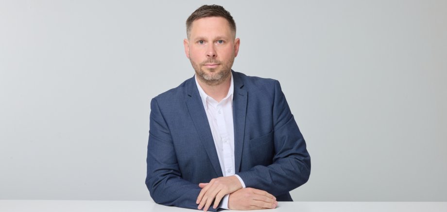 Ein Mann mit Bart und blauem Jackett lehnt an einem Tisch.