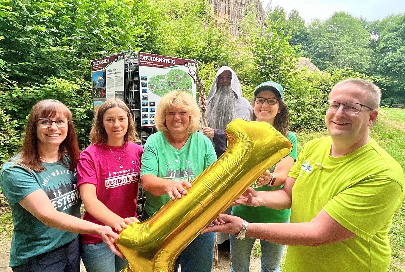 Fünf Personen halten eine goldene 1 in der Hand und stehen mit einem Druiden vor dem Schild des Wanderwegs.