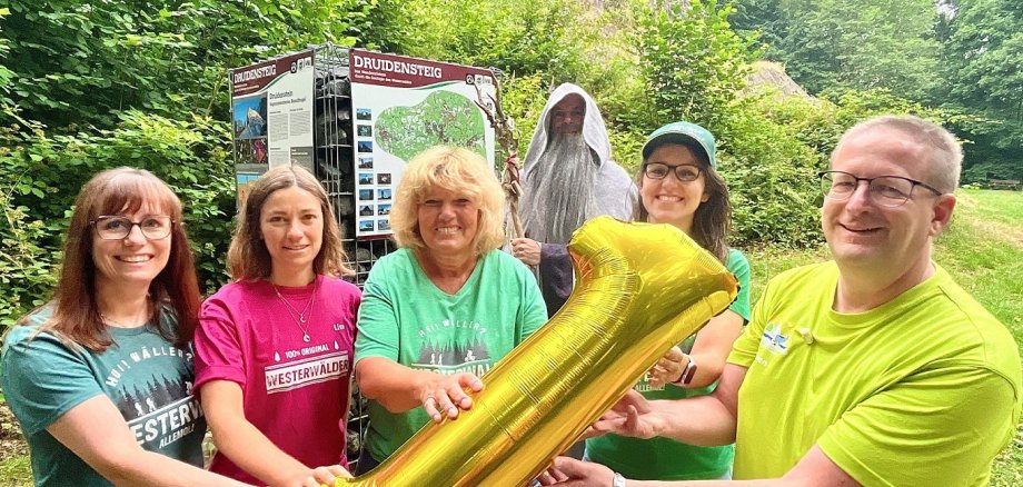 Fünf Personen halten eine goldene 1 in der Hand und stehen mit einem Druiden vor dem Schild des Wanderwegs.