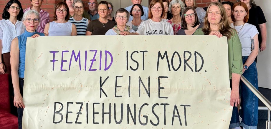 Eine Gruppe von Menschen hält ein Plakat hoch mit der Aufschrift: Femizid ist Mord keine Beziehungstat.