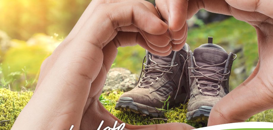 Finger bilden ein Herz um Wanderschuhe, die auf einer Wiese stehen.