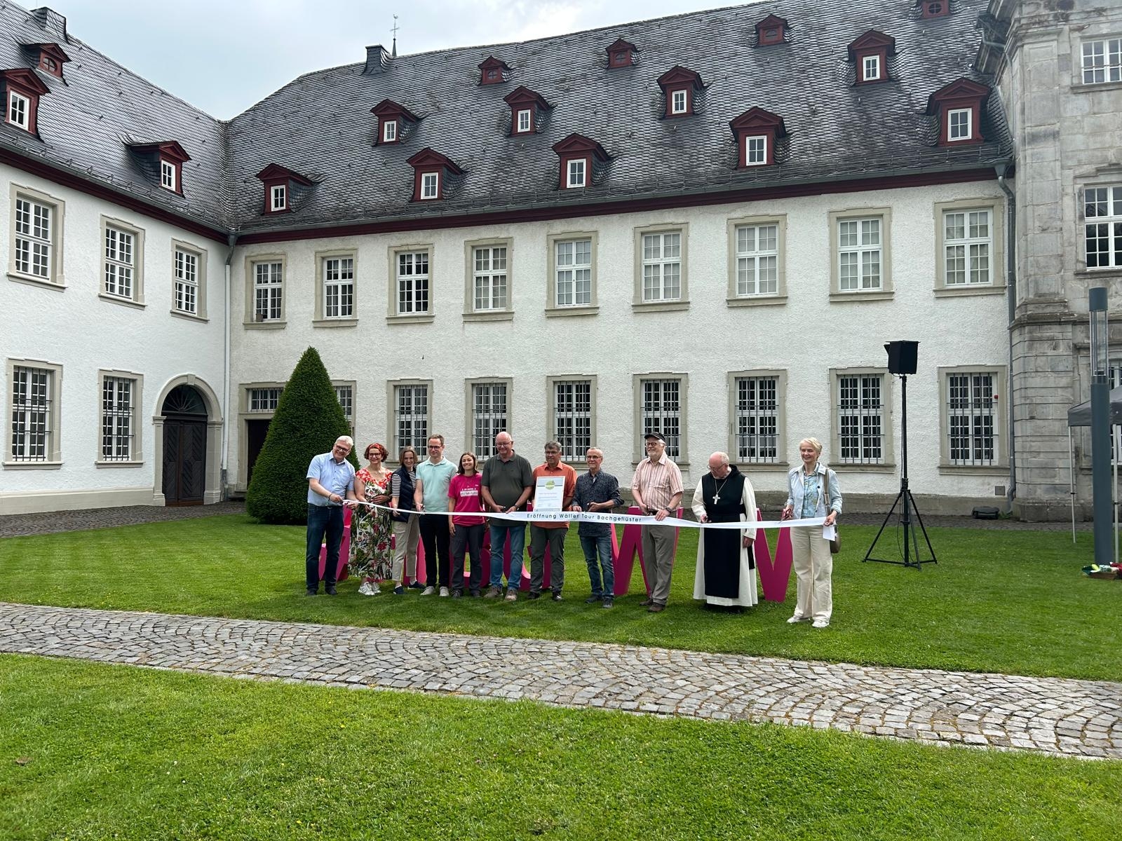 An einem Tische stehen Männer und Frauen vor einem Kloster.
