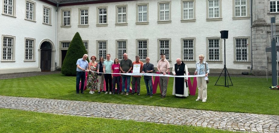 An einem Tische stehen Männer und Frauen vor einem Kloster.