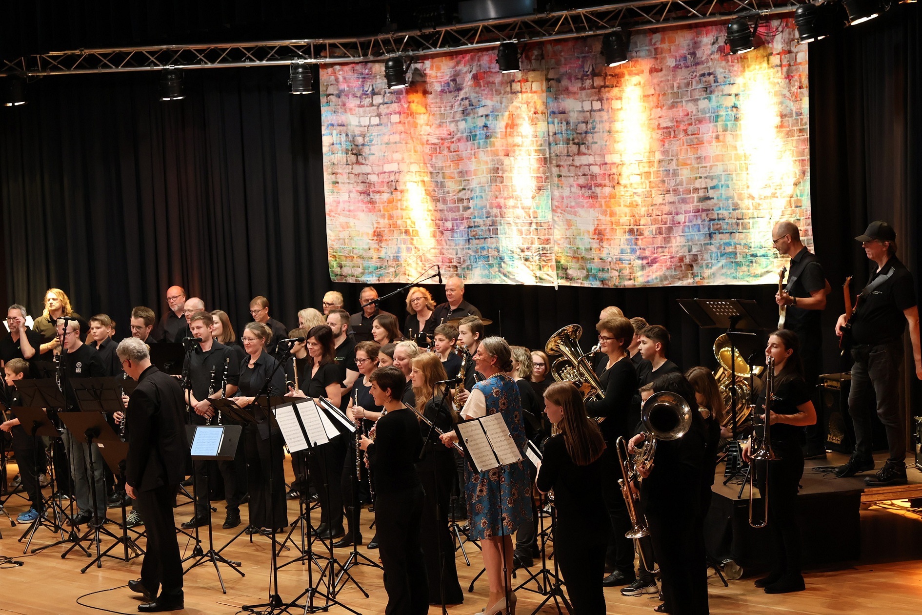 Zahlreiche Musikerinnen und Musiker sind mit verschiedenen Blasinstrumenten auf einer Bühne.