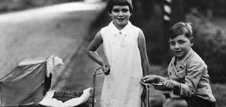 Auf dem alten Schwarz-Weiß-Foto ist ein Mädchen mit Puppenwagen und ein Junge auf einem Dreirad.