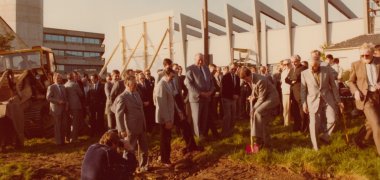 Viele Männer in Anzügen stehen auf einer Wiese, einer rammt einen roten Spaten in den Boden.