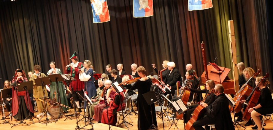 Auf einer großen Bühne sitzen Musiker allen Alters mit verschiedenen Instrumenten.