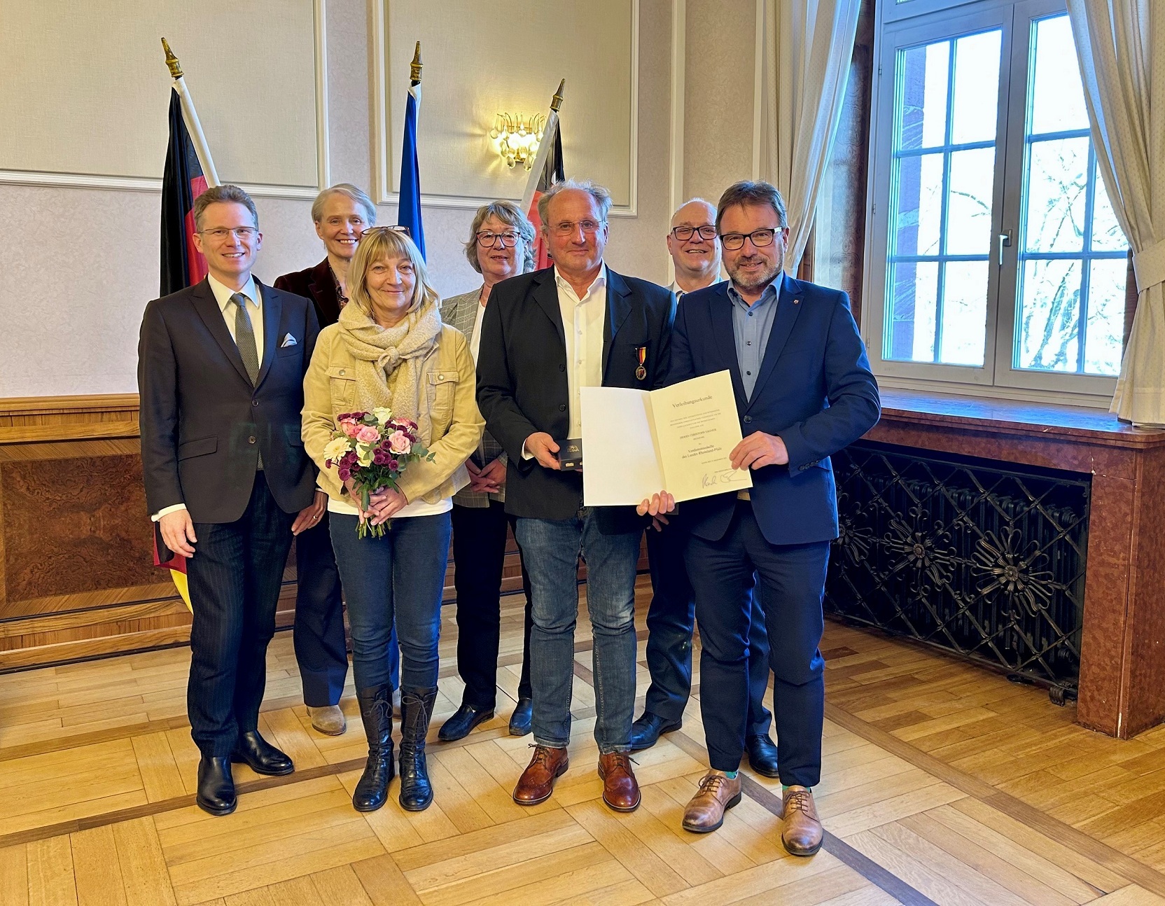 Mehrere Frauen und Männer stehen vor drei Fahnen, einer mit Urkunde und einer mit Medaille.
