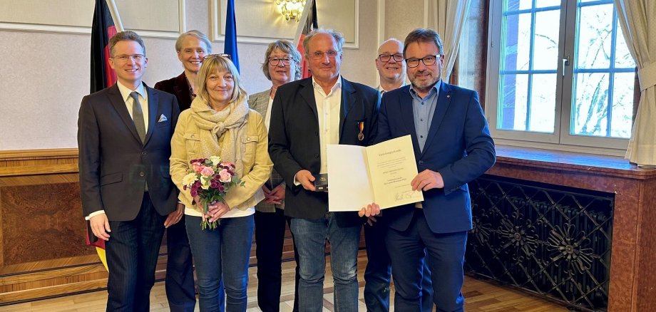 Mehrere Frauen und Männer stehen vor drei Fahnen, einer mit Urkunde und einer mit Medaille.