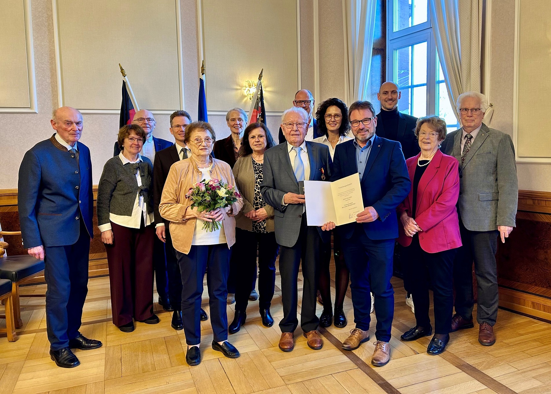 Frauen und Männer stehen vor drei Fahnen, einer mit Medaille und einer mit Urkunde.