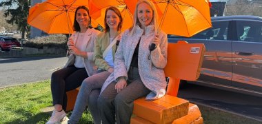Drei Frauen mit orangen Regenschirmen sitzen auf einer orangen Bank.