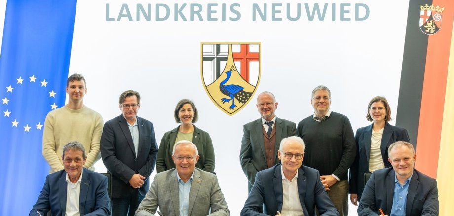 Vier Männer unterzeichnen am Tisch Verträge, dahinter stehen weitere Personen.