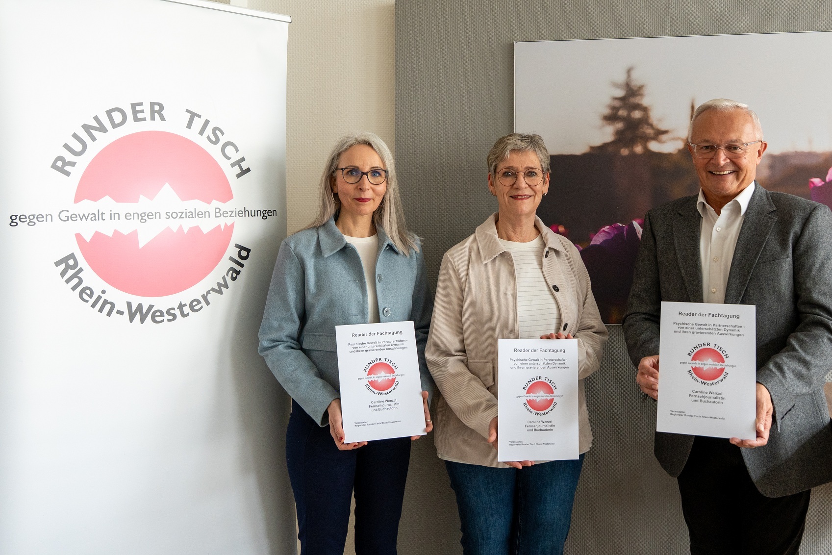 Neben dem Banner des Runden Tisches stehen zwei Frauen und ein Mann mit dem Reader in der Hand.