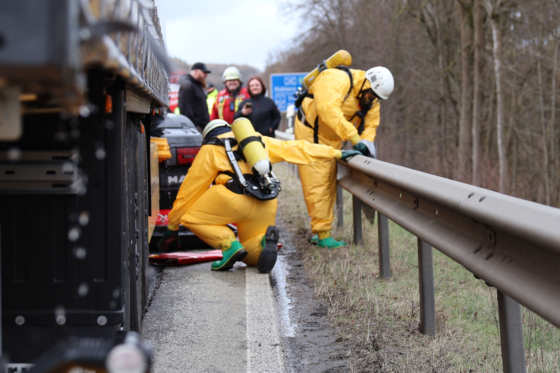 Zwei Personen in gelben Schutzanzügen fangen mit einer Wanne Flüssigkeit neben einem LKW auf.