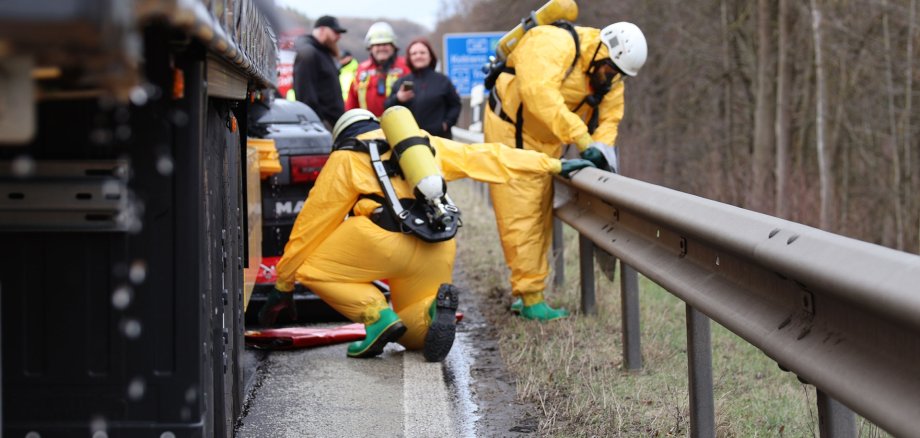 Zwei Personen in gelben Schutzanzügen fangen mit einer Wanne Flüssigkeit neben einem LKW auf.