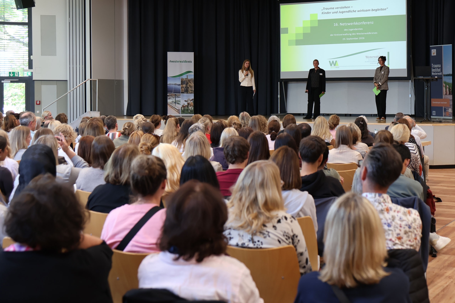 Drei Frauen stehen auf einer Bühne vor vollbesetzten Stuhlreihen.