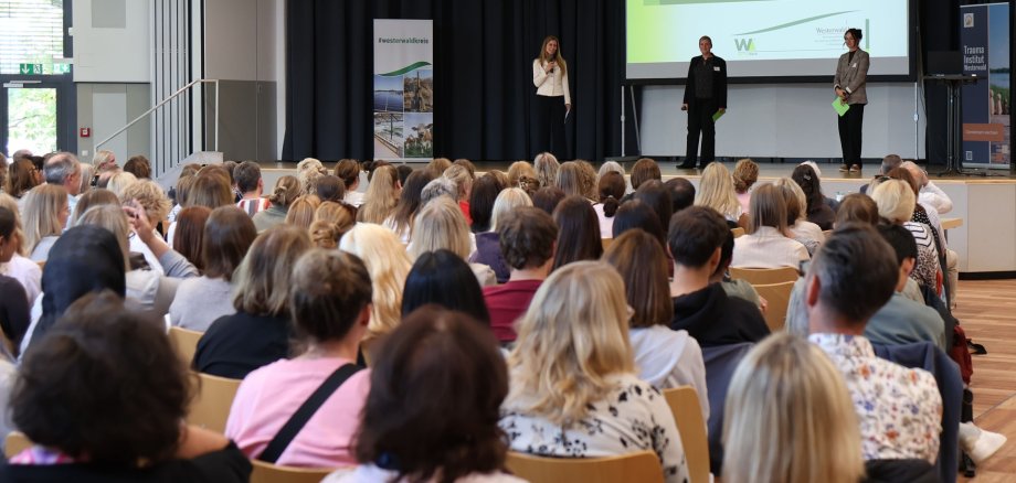Drei Frauen stehen auf einer Bühne vor vollbesetzten Stuhlreihen.