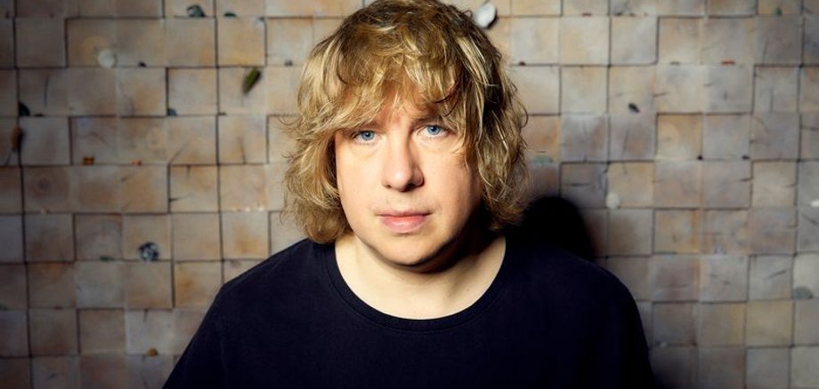 Ein Mann mit lockigem blonden Haar und schwarzem Pullover.