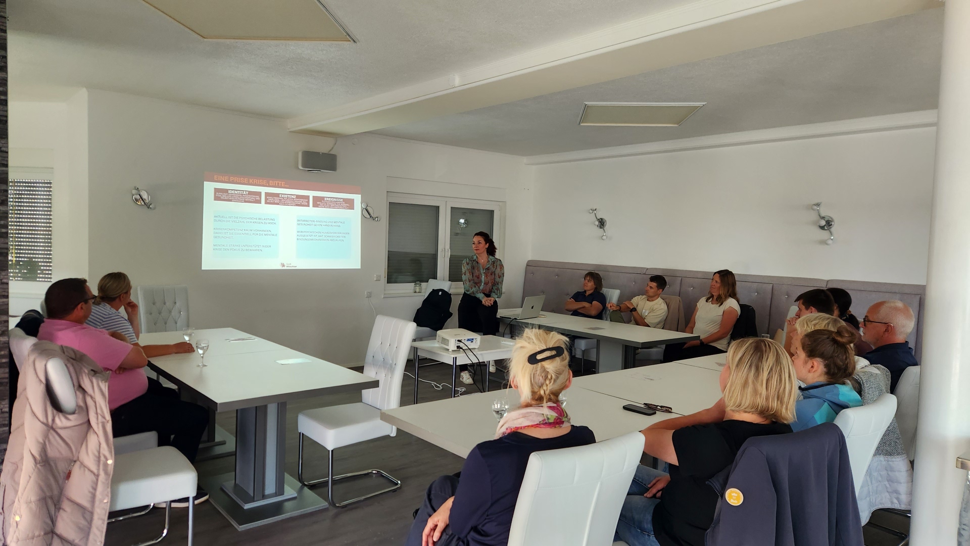 Menschen sitzen an Tischen in U-Form und verfolgen eine Präsentation.