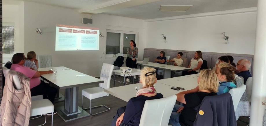 Menschen sitzen an Tischen in U-Form und verfolgen eine Präsentation.