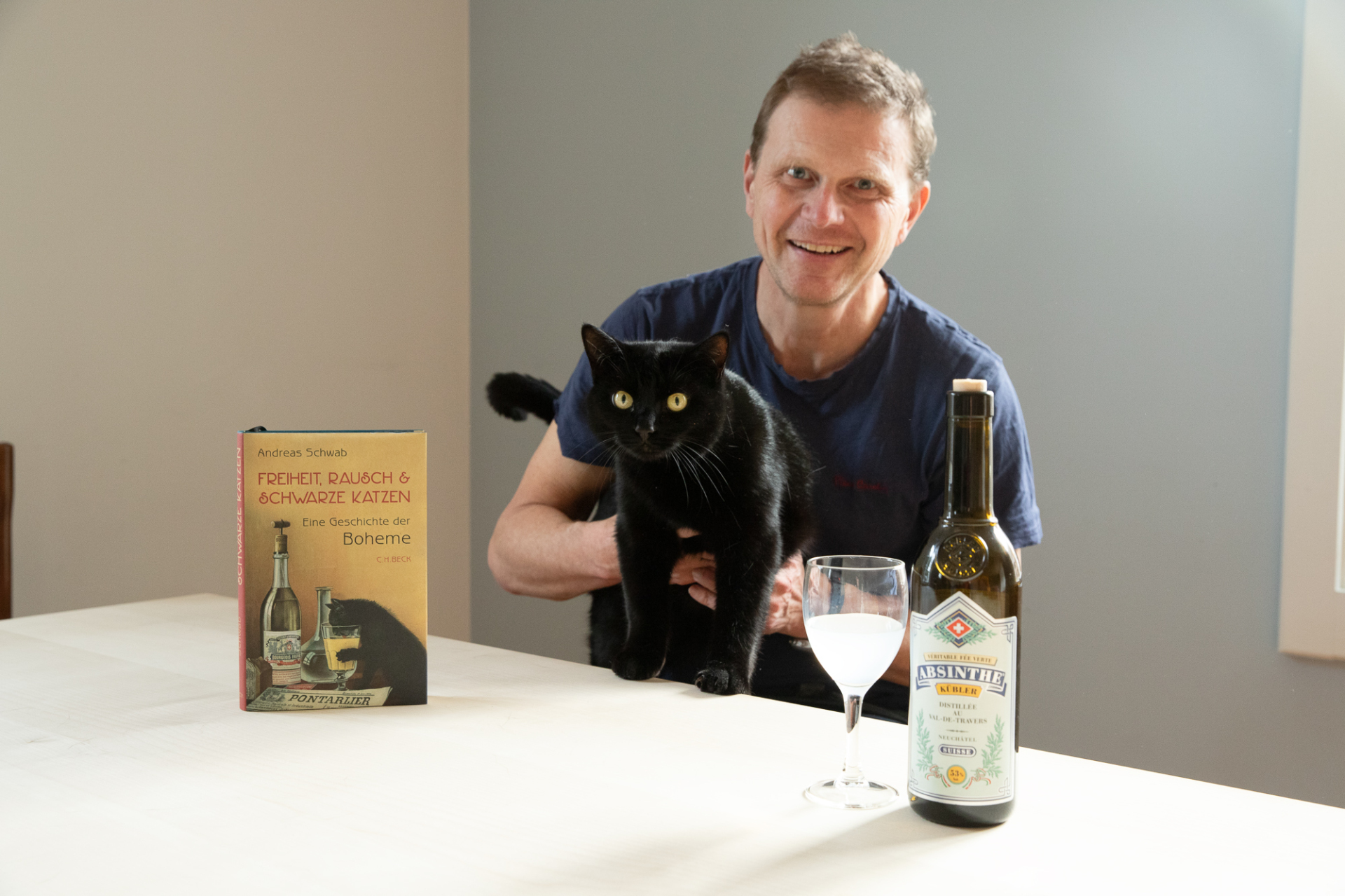 Ein Mann sitzt am Tisch mit Buch, Wein und schwarzer Katze.