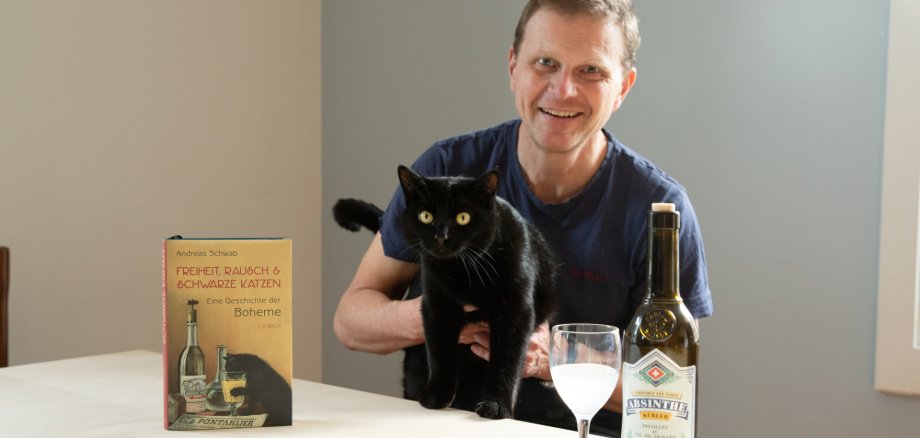 Ein Mann sitzt am Tisch mit Buch, Wein und schwarzer Katze.
