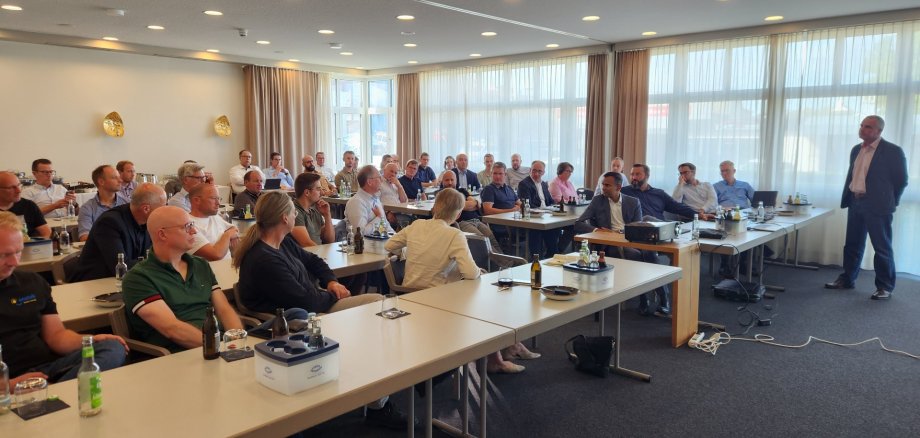 Viele Menschen sitzen in einem Seminarraum an Tischen.