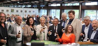 Frauen und Männer stehen mit einer Flasche Bier in der Hand im Sitzungssaal.