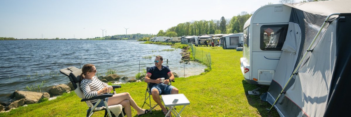 Ein Pärchen frühstückt vor seinem Camper an einem Weiher.
