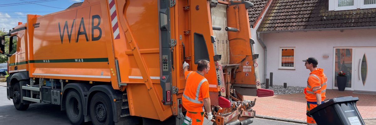 Zwei Mitarbeiter leeren an einem orangenen Müllwagen schwarze Abfalltonnen.