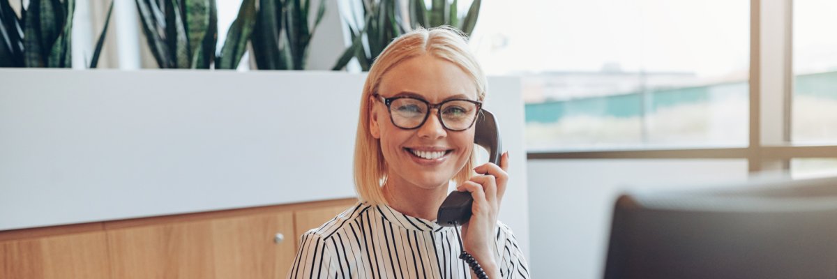 Eine lachende blonde Dame sitzt vor ihrem PC und telefoniert.