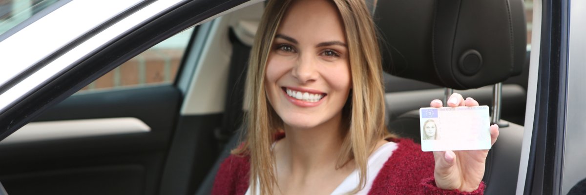 Eine Frau hält lachend ihren Führerschein aus dem Fenster ihres Autos heraus in die Kamera.
