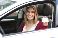Eine junge Frau zeigt im Auto sitzend durch Fahrerfenster lachend ihren Führerschein.