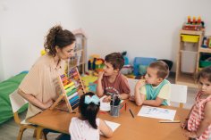 Eine Kindergärtnerin erklärt an einem Tisch sitzend Kindern das Rechnen.
