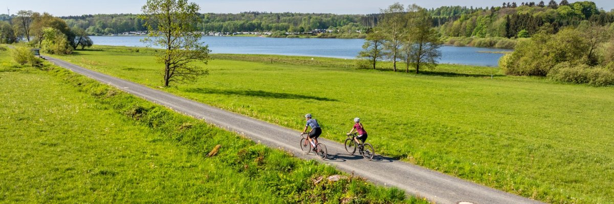 Zwei Radfahrer fahren entlang der Krombachtalsperre durch grüne Wiesen.