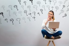 Eine junge Frau im Schneidersitz mit Laptop auf dem Schoss und fragender Gestik.