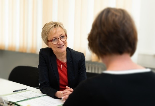 Eine Frau mit kurzem blonden Haar, Brille und Jackett sitzt an einem Schreibtisch im Gespräch mit einer anderen Frau.