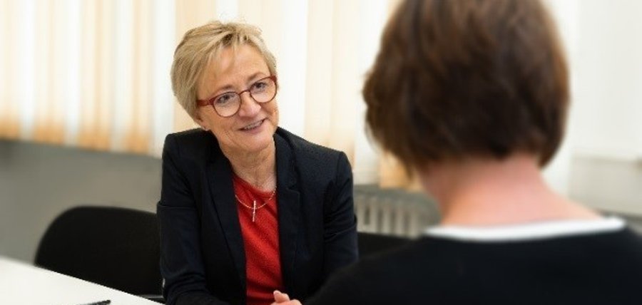 Eine Frau mit kurzem blonden Haar, Brille und Jackett sitzt an einem Schreibtisch im Gespräch mit einer anderen Frau.