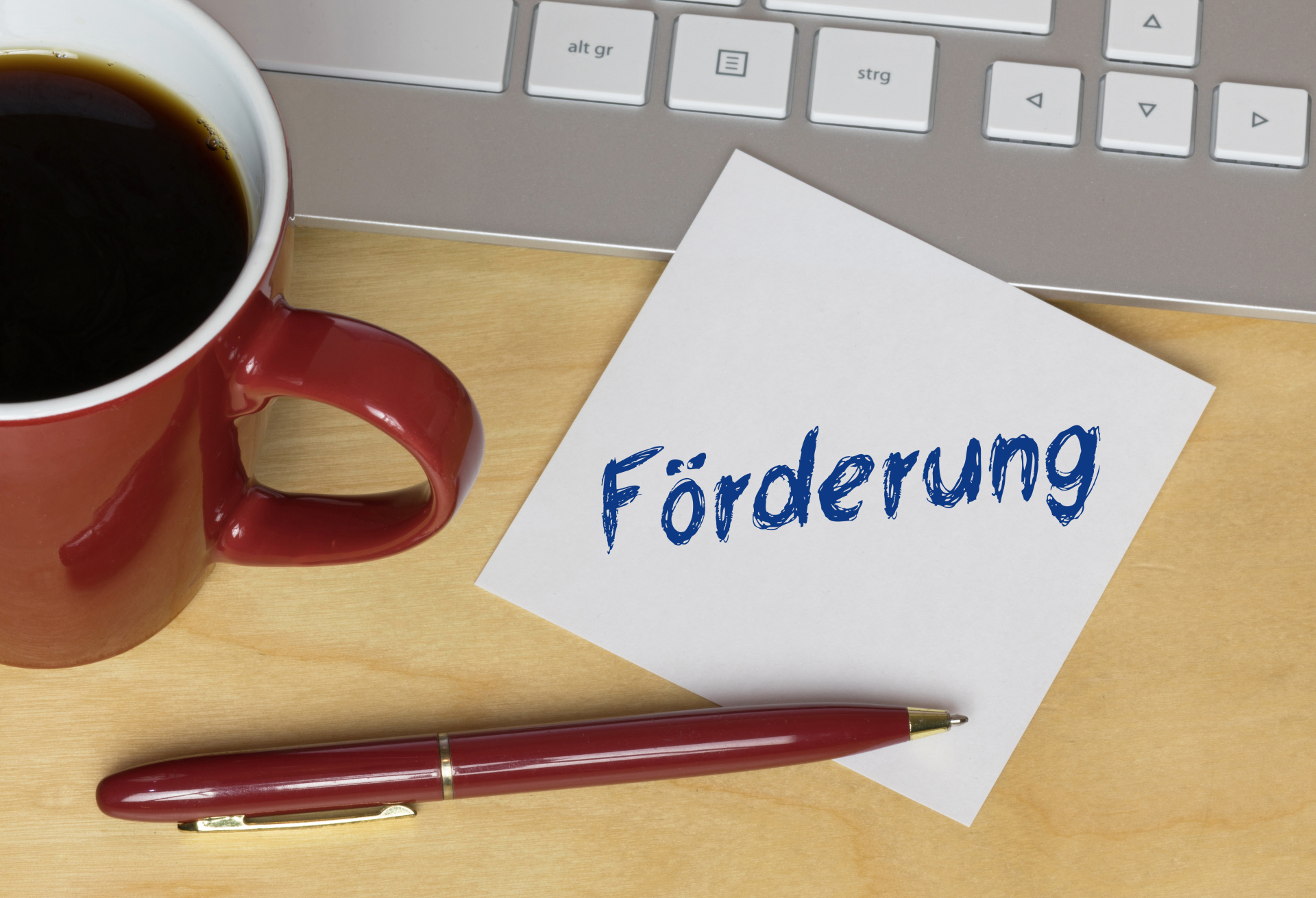 Auf einem Schreibtisch steht eine volle rote Kaffeetasse, ein Laptop und ein Zettel auf dem Förderung steht.