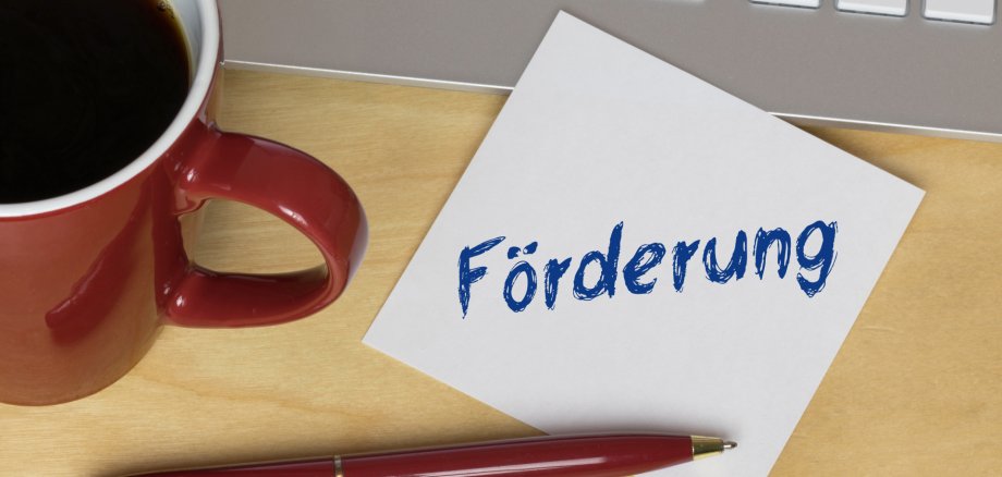 Auf einem Schreibtisch steht eine volle rote Kaffeetasse, ein Laptop und ein Zettel auf dem Förderung steht.