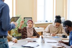 Eine muslimische Frau mit Kopftuch fragt ihren Lehrer während einer Integrationsstunde etwas. Eine weitere Frau und ein Mann sitzen mit am Tisch und lesen in Büchern.