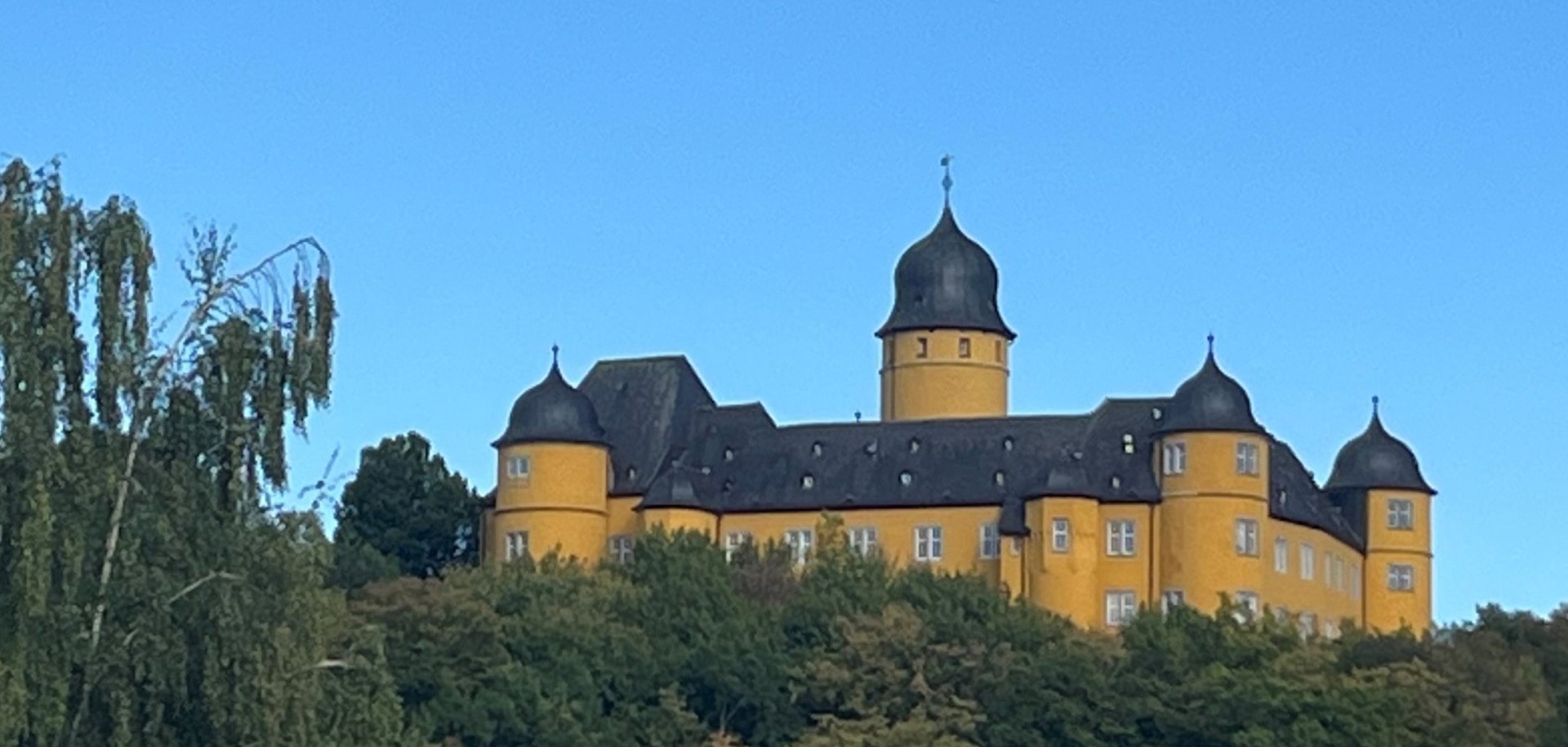 Ein gelbes Schluss steht inmitten von Bäumen auf einem Hügel vor blauem Himmel.