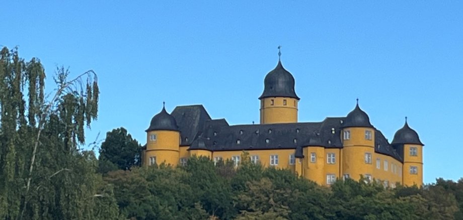 Ein gelbes Schluss steht inmitten von Bäumen auf einem Hügel vor blauem Himmel.