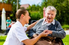 Eine Krankenschwester hält die Hand einer lachenden Seniorin, die im Rollstuhl in der Sonne im Garten sitzt.