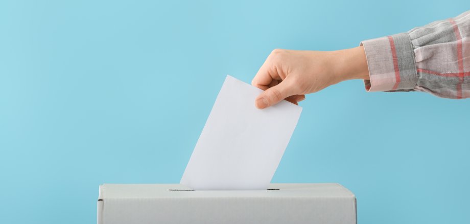 Eine Hand steckt einen Wahlzettel in eine Urne.